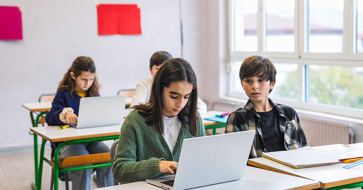 Four students sit at desks in a classroom, working on laptops and notebooks with AI-powered tools, as windows and colorful papers adorn the wall in the background.