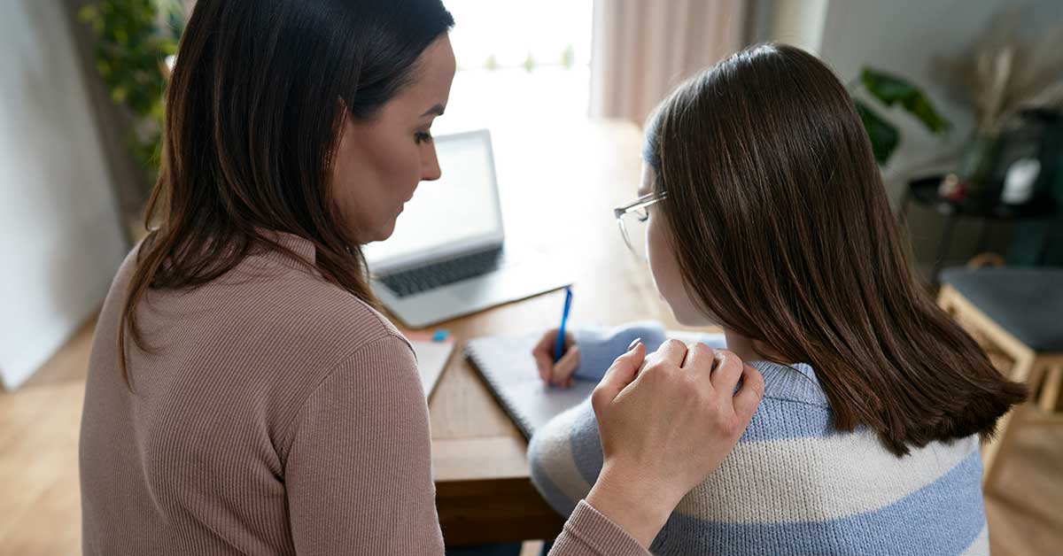 A woman sits beside a girl who is writing in a notebook at a table with a laptop, resting her hand supportively on the girl's shoulder as they discuss the dangers of sextortion and how to stay safe online.