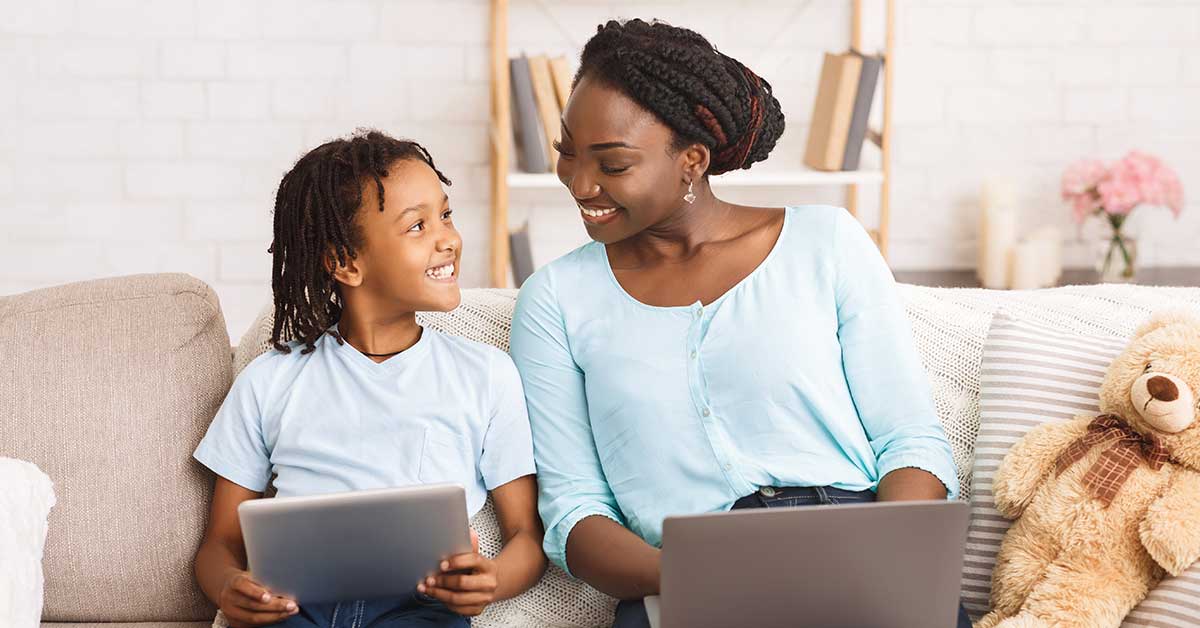 An adult and child sit on a couch, smiling at each other. The child holds a tablet exploring AI in education while the adult uses a laptop. A teddy bear is next to them.