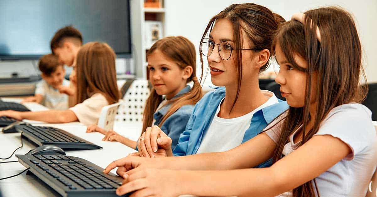 A teacher assists a young girl at a computer while other children use AI-powered tools at computers in a classroom setting.