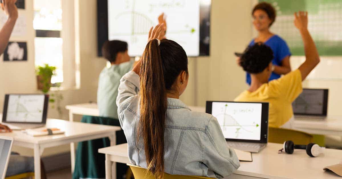 Students in a classroom raise their hands while seated at desks with laptops, engaging in a lesson on AI as the teacher stands near a screen displaying a graph.