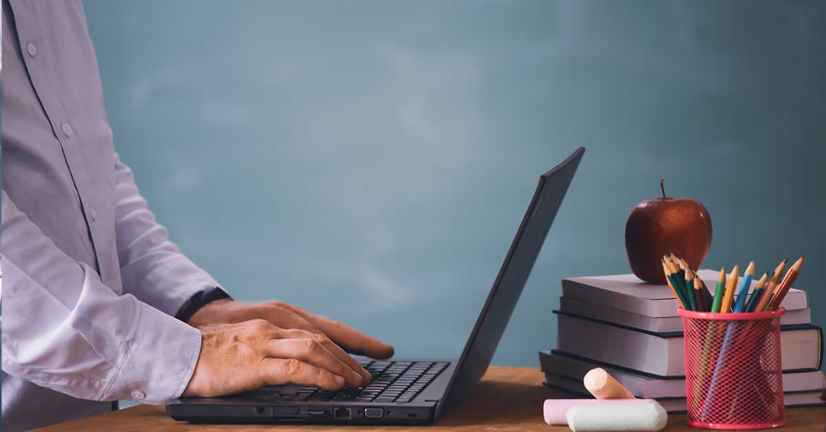 Person typing on a laptop at a desk with stacked books, an apple, chalk, and a cup of pencils in front of a chalkboard, focused on organizing student data.