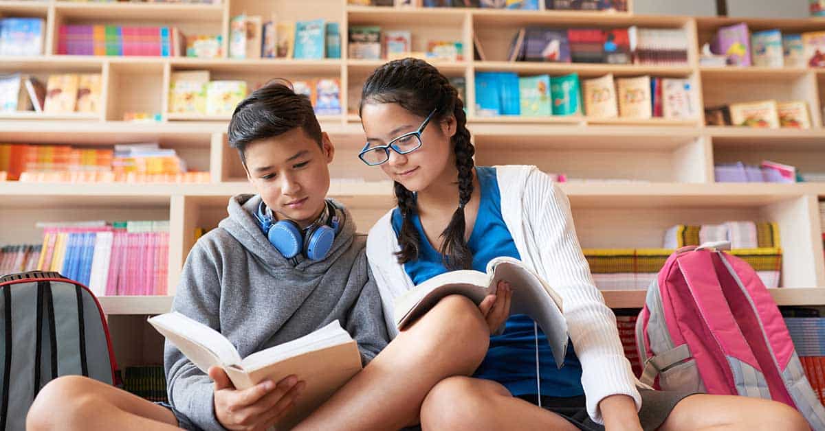 Two students sit on the floor in a library, fostering literacy as they read books together, with backpacks beside them and bookshelves in the background.