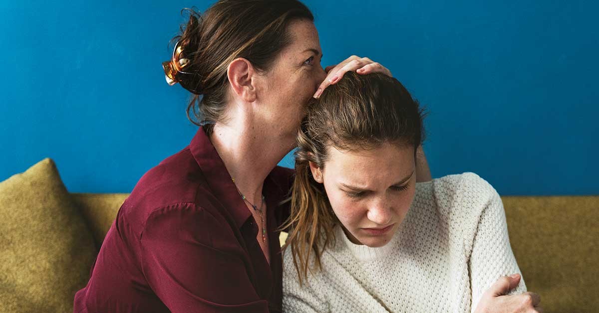 A woman comforts a visibly upset young girl on a couch, holding her close and gently touching her head, offering support after the girl shared her fears about sextortion.
