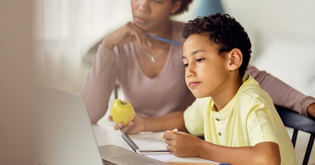 A young boy sits at a table doing schoolwork on a laptop, guided by an adult holding an apple and watching attentively—a helpful scene from a guide for parents supporting their children’s learning.