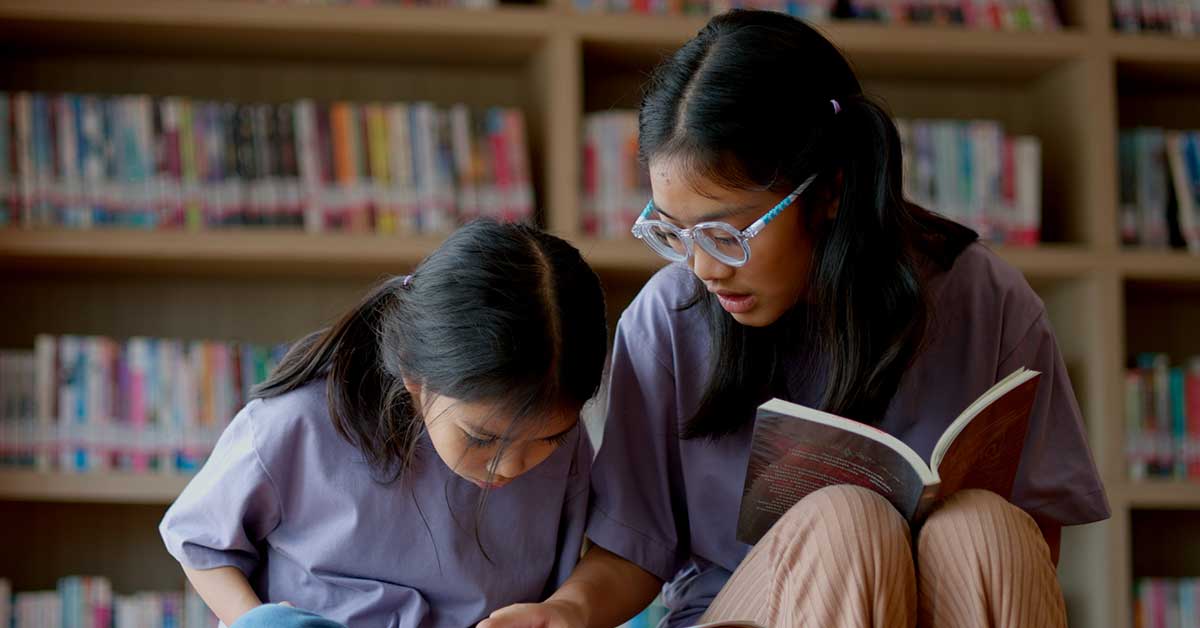 Two girls sit together in a library, one reading a book aloud while the other follows along, building their reading proficiency. Bookshelves are visible in the background.