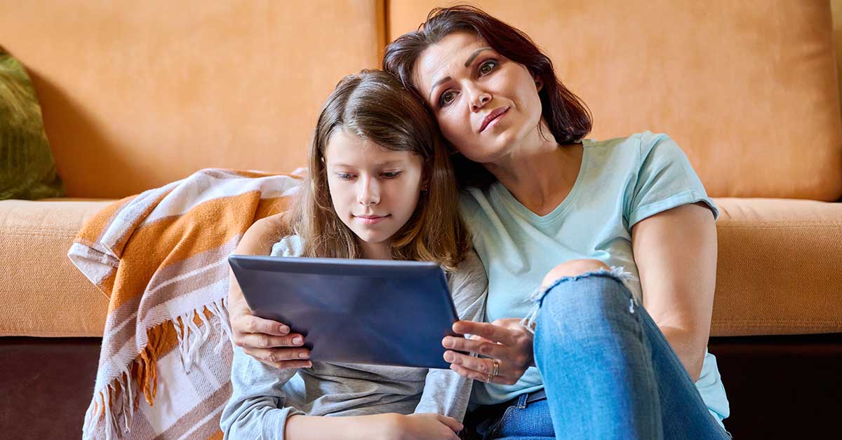 An adult and a child sit on the floor, leaning against a couch and looking at a tablet together, highlighting the importance of internet security for family safety.