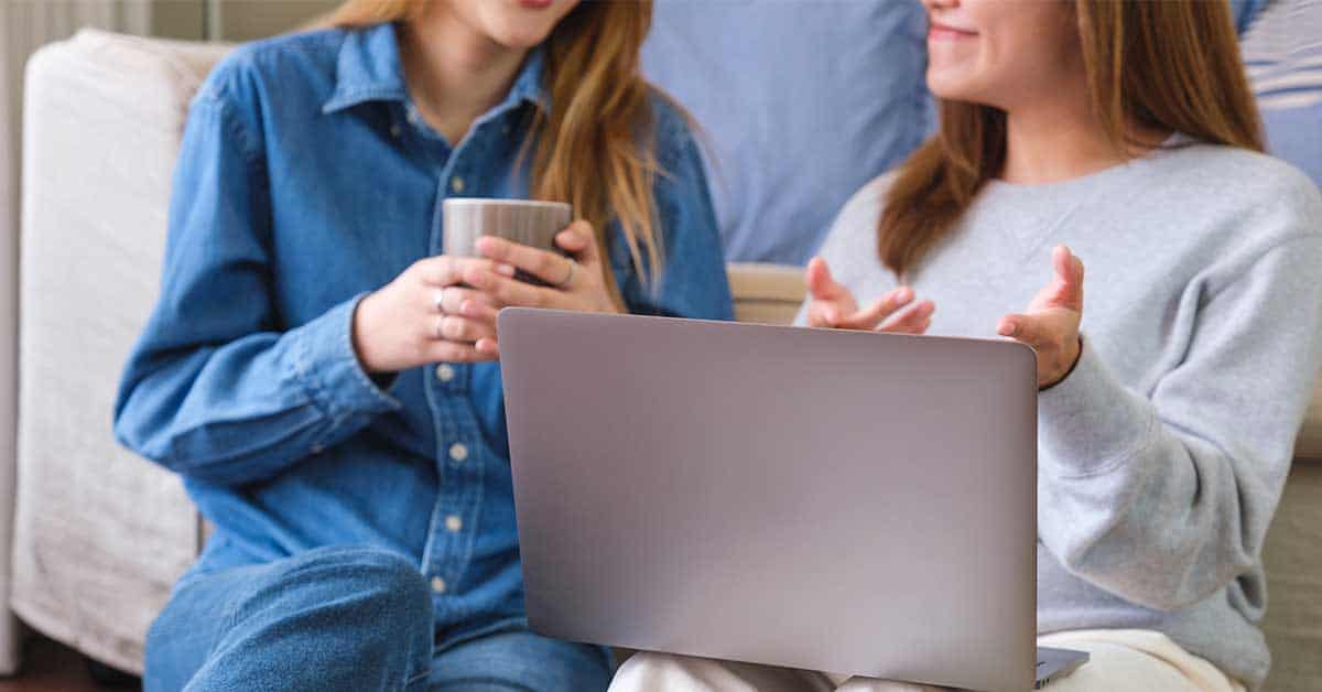 Two people sitting indoors, one holding a mug and the other gesturing with hands, discuss sextortion with an open laptop on their laps.