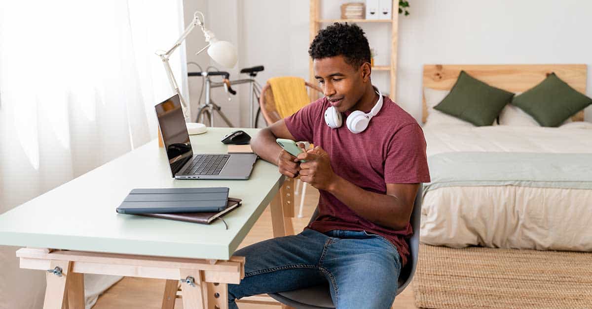 A young man sits at a desk in his bedroom, looking at his phone, with a laptop, tablet, and headphones—tools that highlight the digital divide faced by many underserved school communities seeking effective strategies for learning.