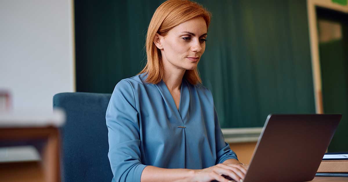 A woman with red hair wearing a blue blouse sits at a desk in a classroom setting, typing on a laptop as she explores AI applications.