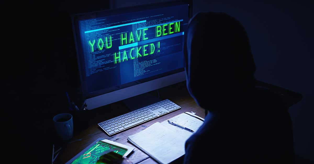 Person in hoodie at desk facing computer screen with “You have been hacked!” in green text amid code, highlighting account security risks in a dark room.