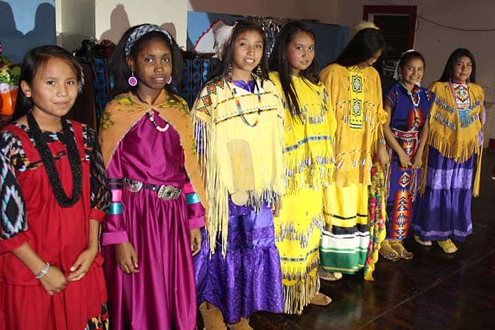 A group of young girls stand in a row wearing colorful traditional Native American dresses and accessories, indoors on a wooden floor.