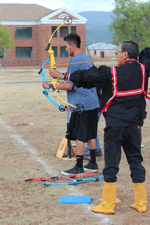 Two people stand in a field aiming compound bows at a target off camera; archery equipment is on the ground beside them. A brick building and trees are in the background.