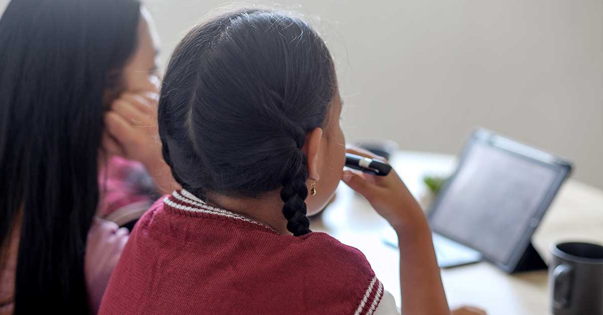 Two people sit at a table, looking at a tablet screen, practicing digital literacy. One holds a pen to her mouth, and both have dark hair tied back. Cups and a potted plant are on the table.