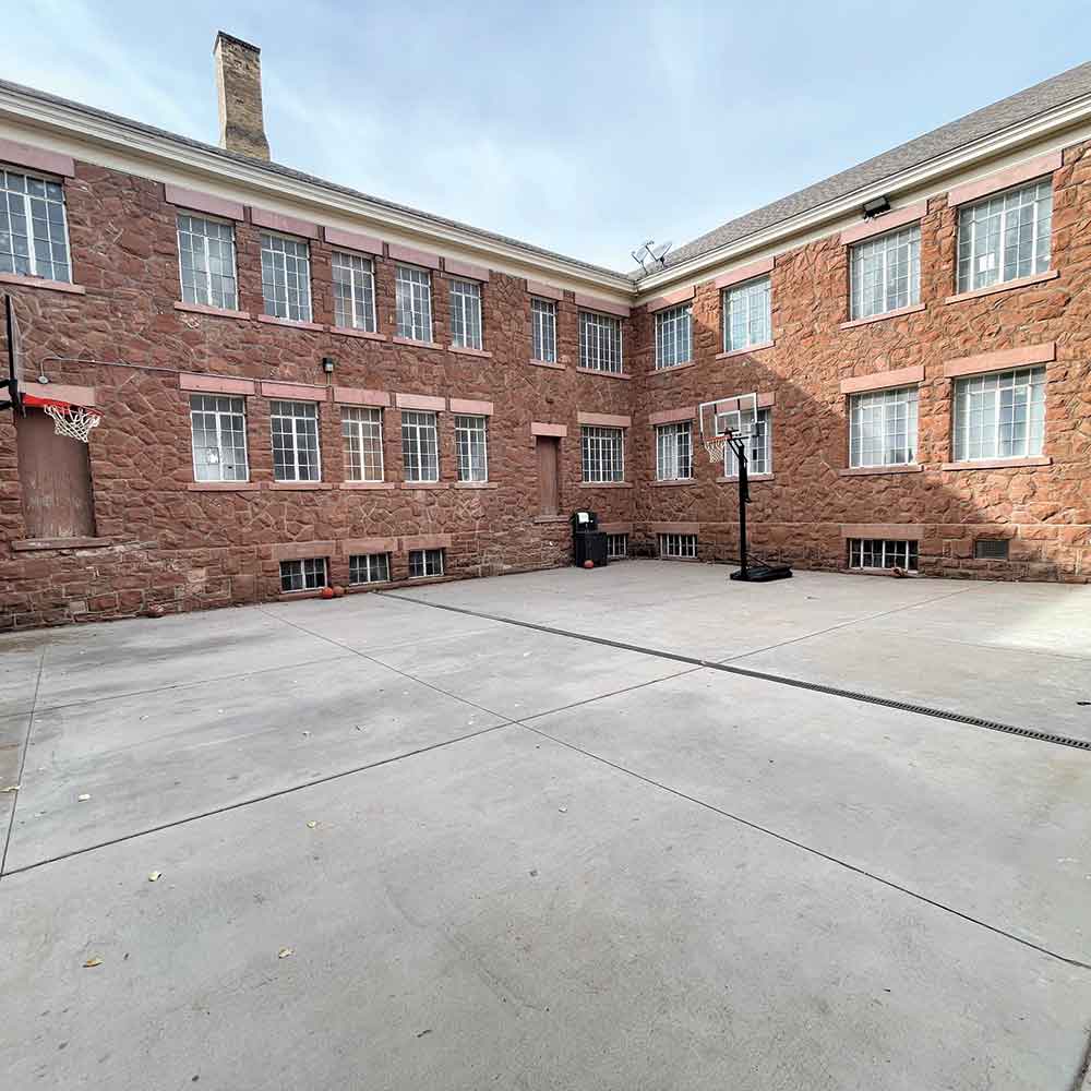 A paved courtyard is surrounded by a two-story red brick building, featuring two basketball hoops and multiple windows.