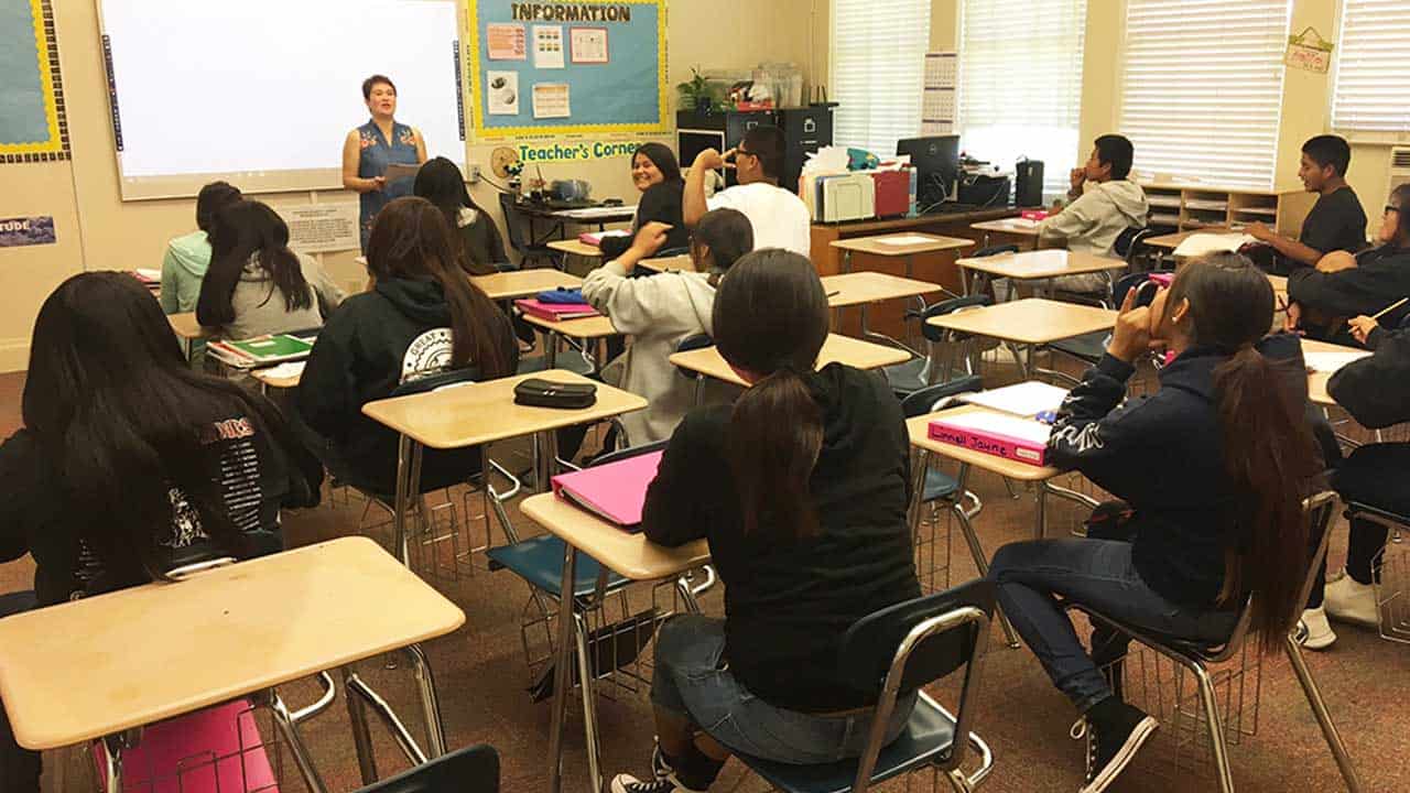 On the First Day of School, a teacher stands at the front of a classroom speaking to students seated at desks, most facing forward.