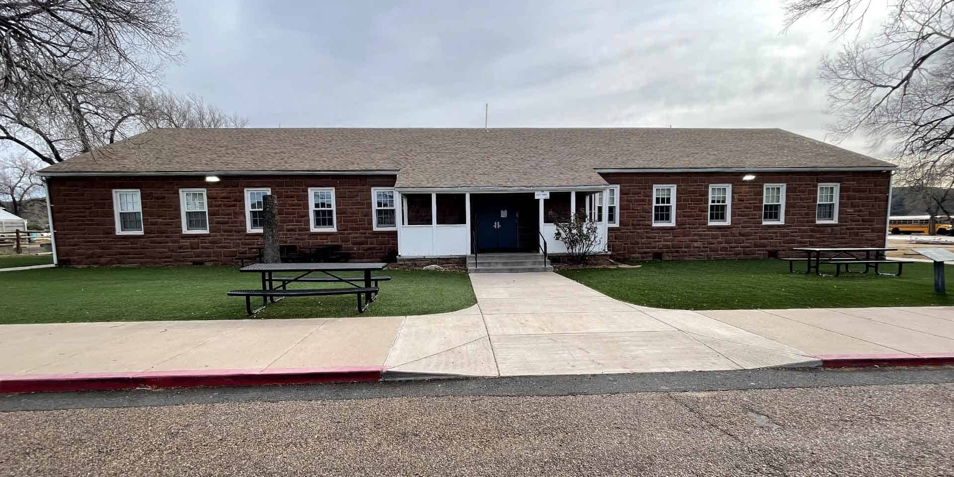 Single-story brick building with a central entrance, multiple windows, picnic tables on either side, and a sidewalk leading to the front door. Trees and grass are visible around the building.