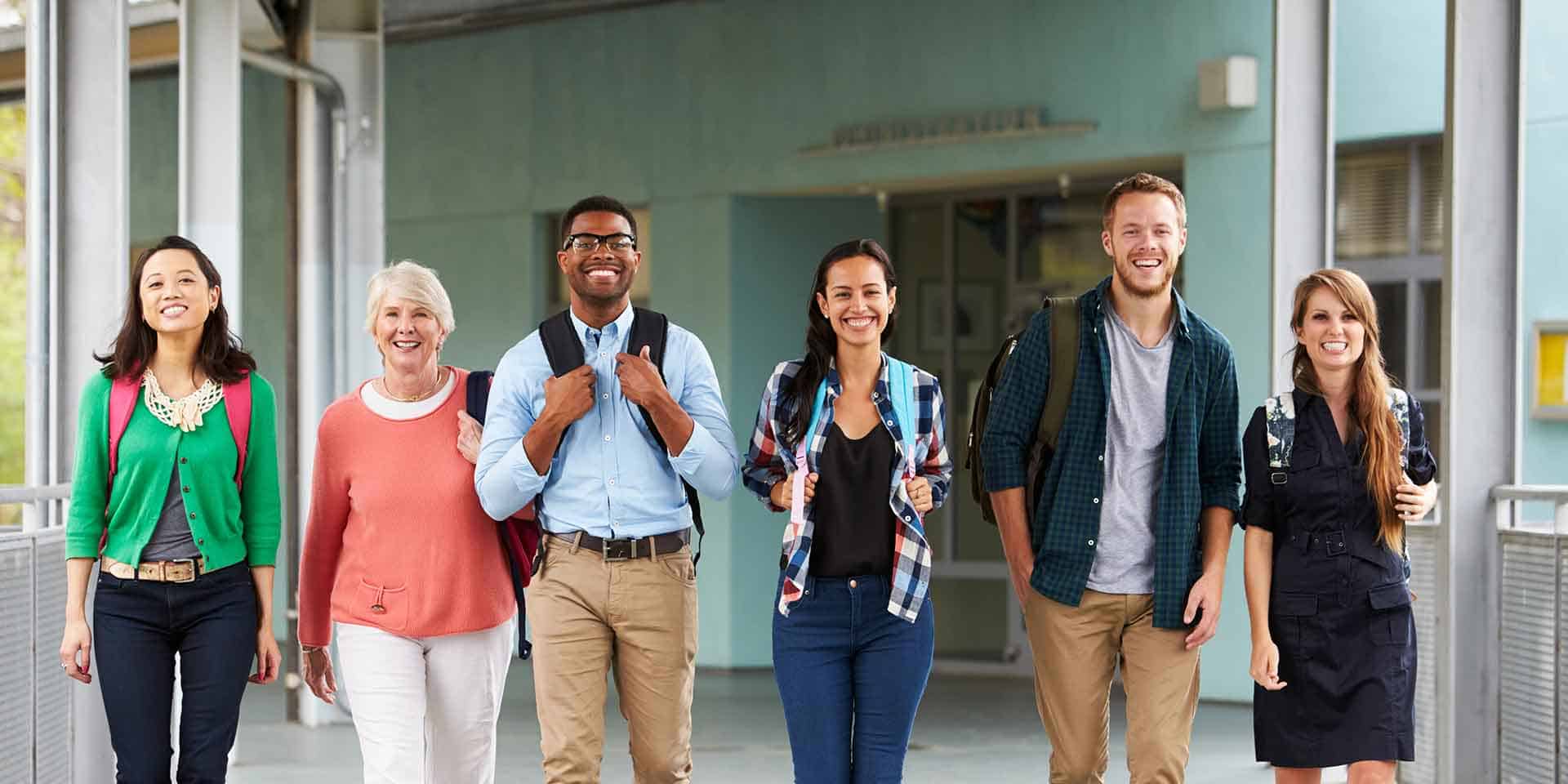 Six people, diverse in age and ethnicity, walk side by side in a hallway, smiling and carrying backpacks.