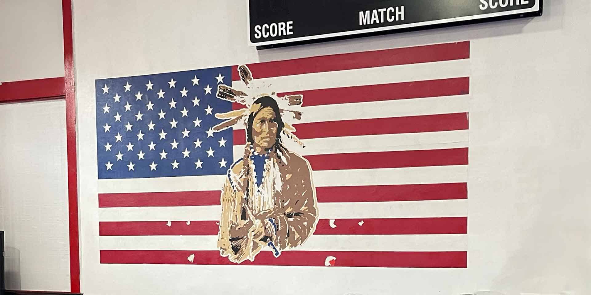 Mural of a Native American man in traditional clothing superimposed on a U.S. flag, painted on an indoor wall beneath a scoreboard.