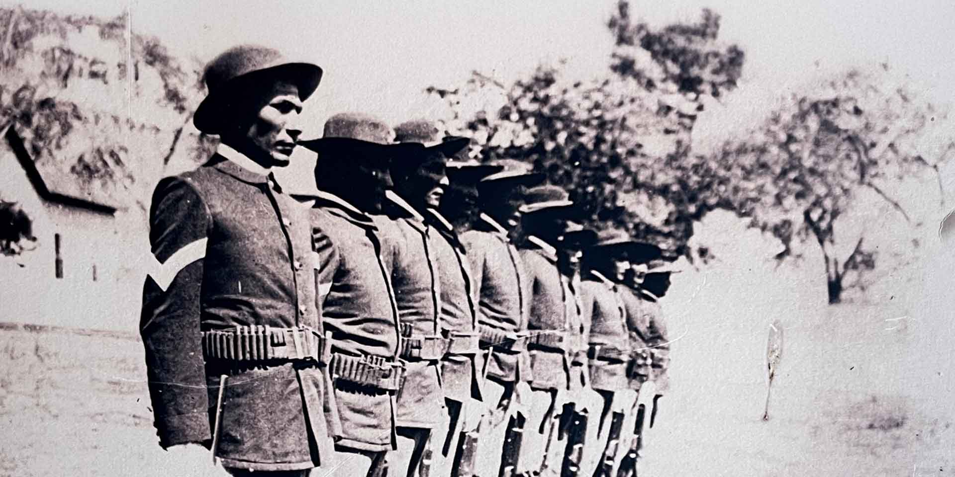 A row of soldiers in uniform and hats stand at attention outdoors, each wearing ammunition belts and facing forward.