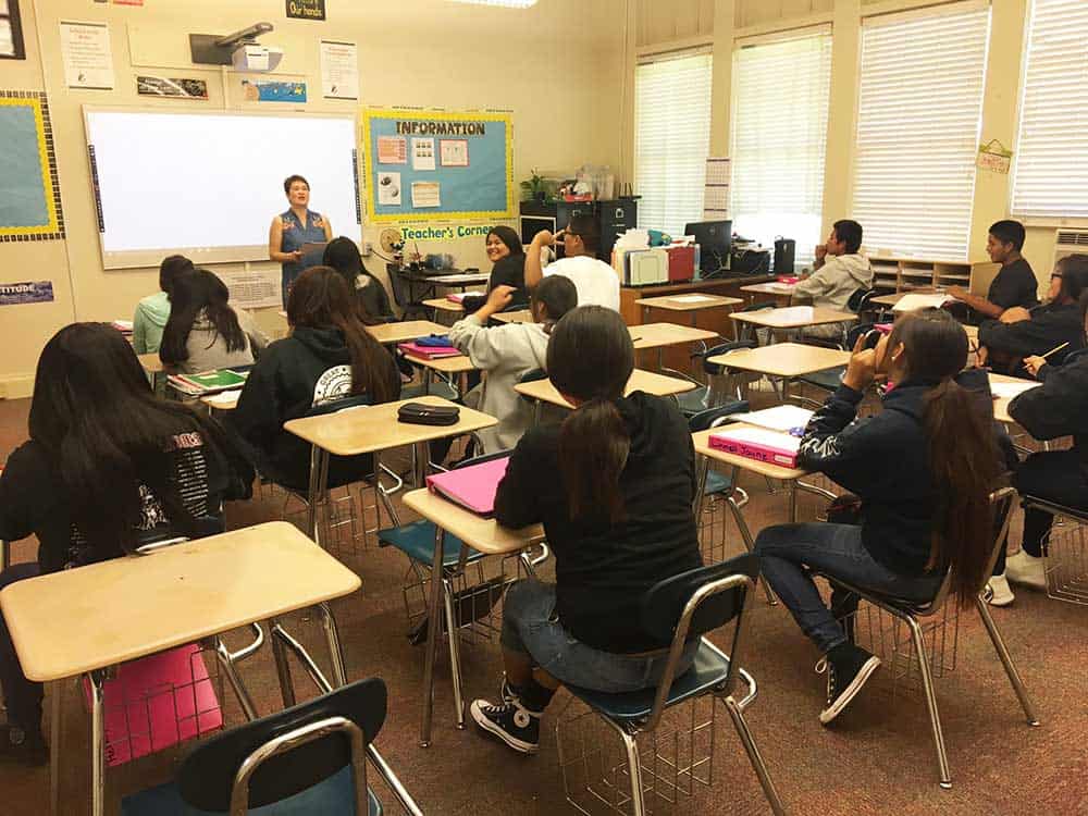 A teacher stands at the front of a classroom speaking to students seated at desks with notebooks and folders.