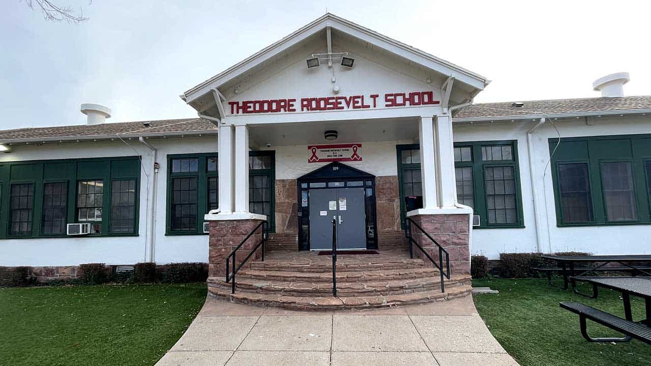 Front view of Theodore Roosevelt School building with white columns, green shutters, and a stone staircase leading to a navy blue door. Benches are visible on the right side.